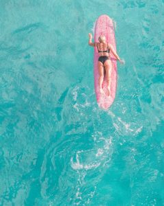 Aerial view of a surfer relaxing on a pink surfboard in clear Hawaiian waters.